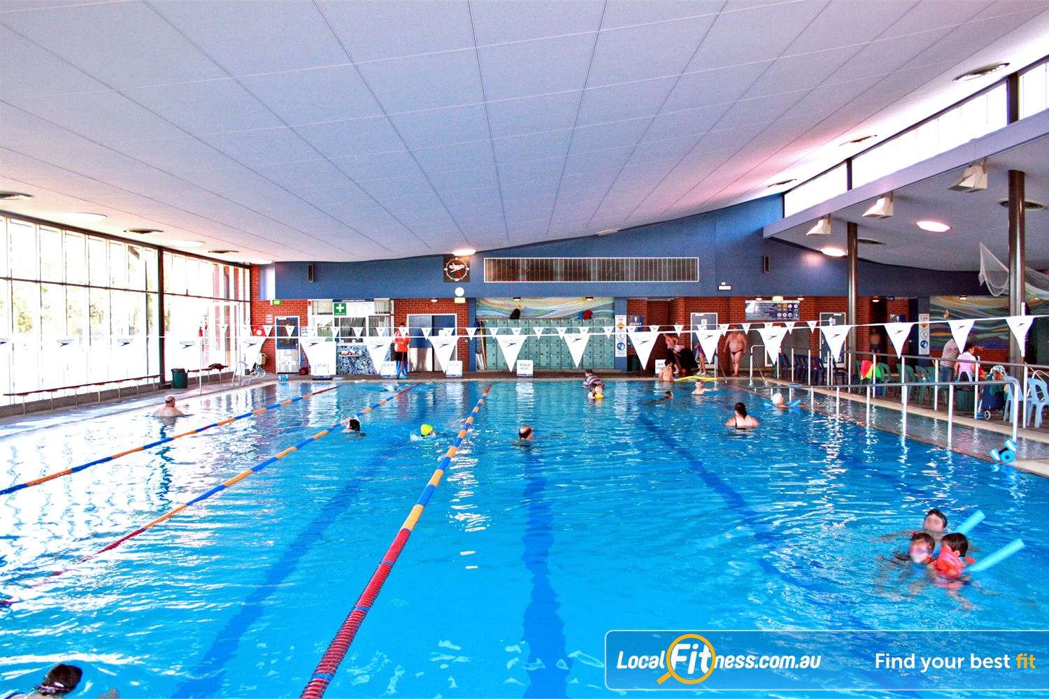 Indoor swimming pool with clear blue water, swimmers of all ages enjoying the space, surrounded by large windows and colorful banners.