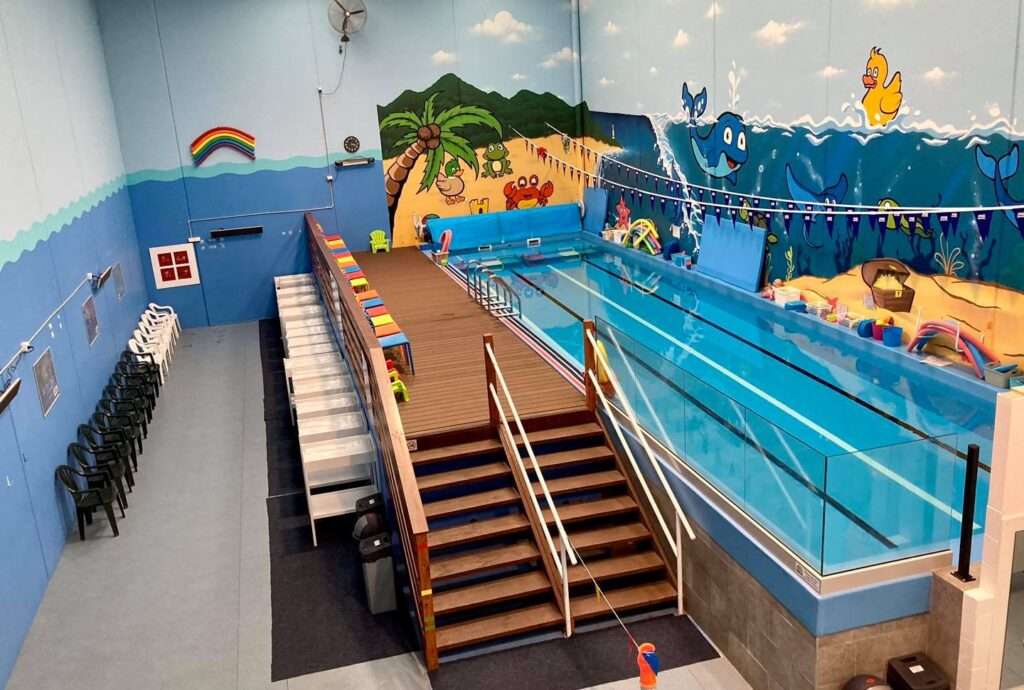 Indoor swimming pool with colorful beach mural, stairs leading to the poolside, and rows of chairs along the wall.