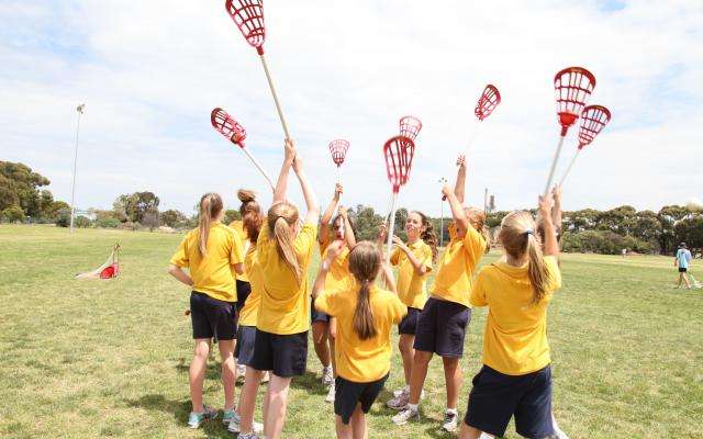 A group of young female lacrosse players in yellow uniforms celebrate with their sticks raised on a sunny field.