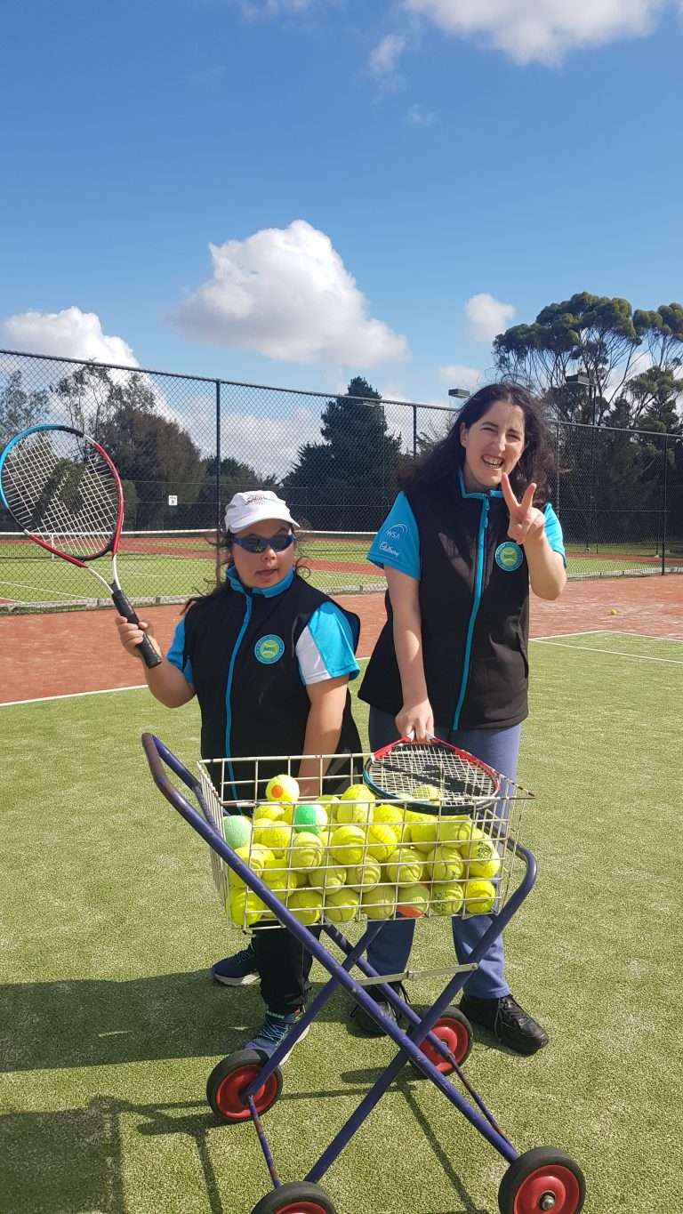 Two smiling individuals on a tennis court, one holding a racket and the other beside a cart filled with tennis balls.