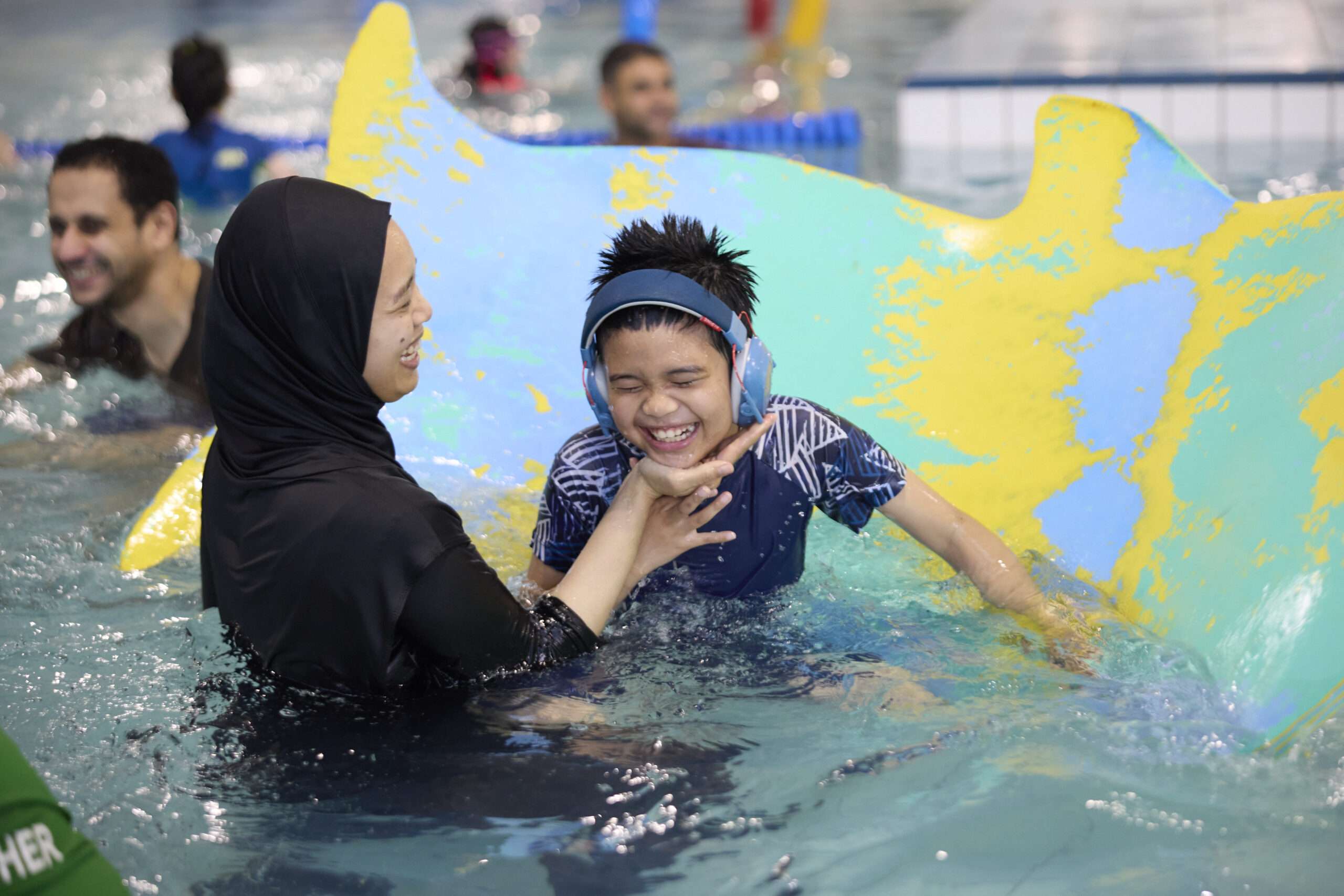 Photo of a group of swimmers in the pool. Photo includes a women holding a young swimmer with headphones on, as she practices her time in the pool.