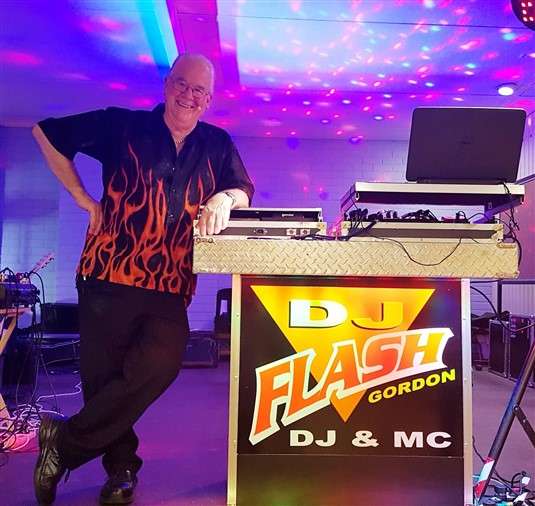 A DJ with a flaming shirt stands beside his equipment at a vibrant event, showcasing his "DJ Flash Gordon" branding.