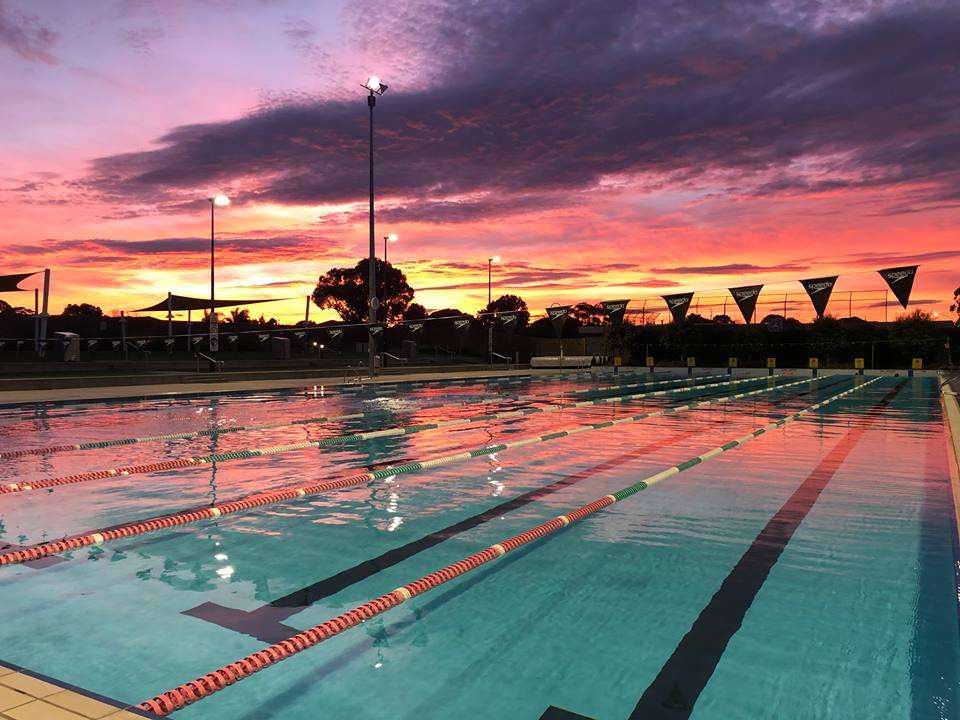 A swimming pool at sunset with vibrant pink and orange skies reflected in the water, lined with lane markers and flags.