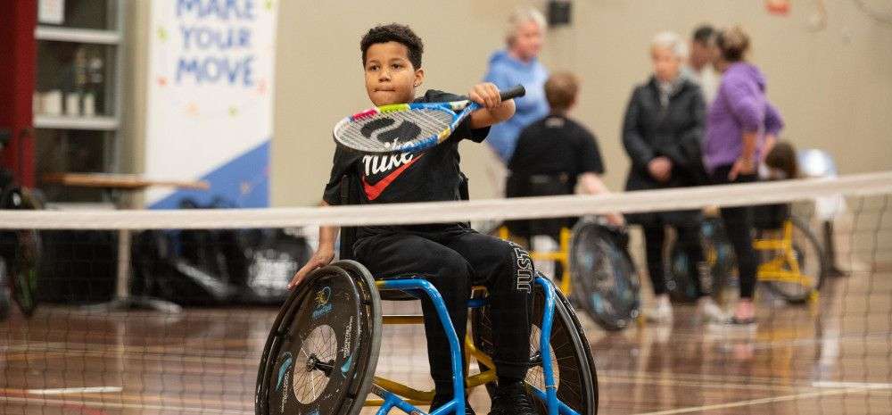 A child in a wheelchair practices tennis, focused on their swing, with others participating in the background of a gym.
