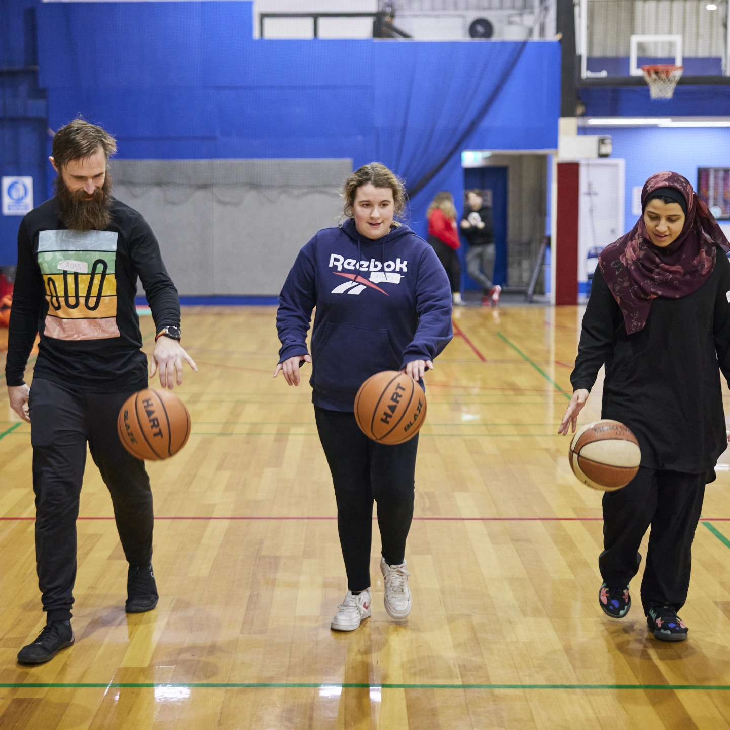 Three people, a man and two women, practice dribbling basketballs on a court, showcasing diversity and teamwork in sports.