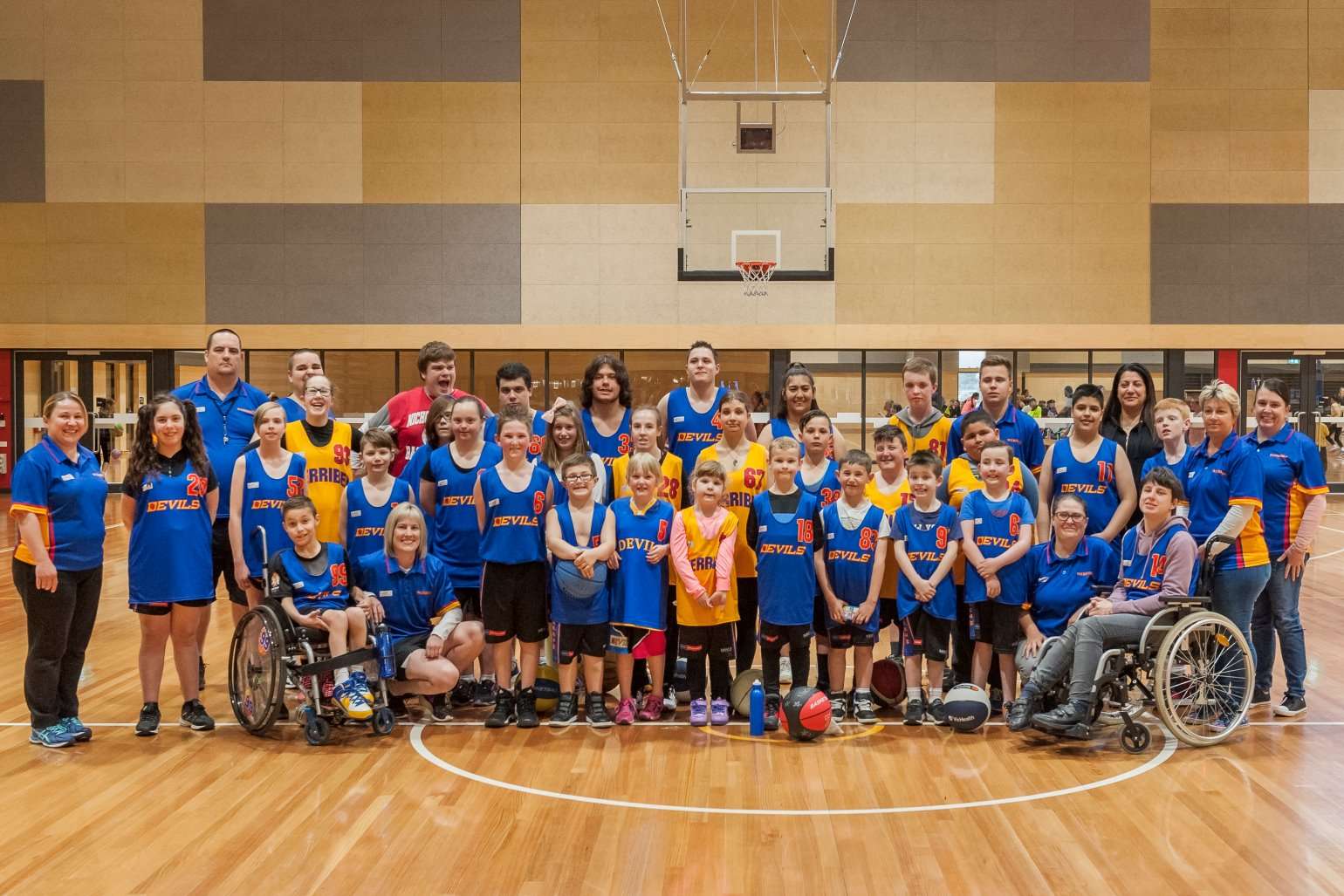 A diverse group of participants in blue and yellow basketball uniforms, gathered in a gym with a basketball hoop in the background.