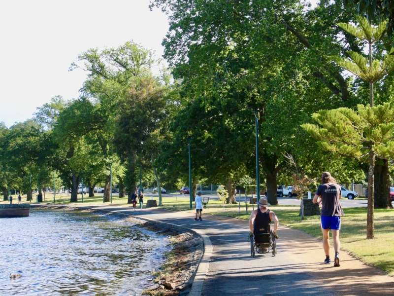 A scenic park pathway along a river, featuring a person in a wheelchair and another individual walking, surrounded by lush greenery.