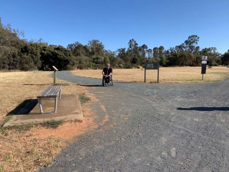 A person in a wheelchair navigates a pathway with a bench and signs in a grassy, open area under a clear blue sky.