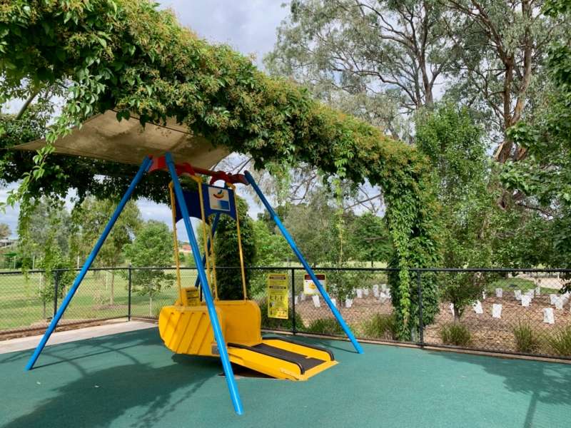 A yellow all abilities swing shaded by foliage, in a fenced playground surrounded by lush greenery and trees.