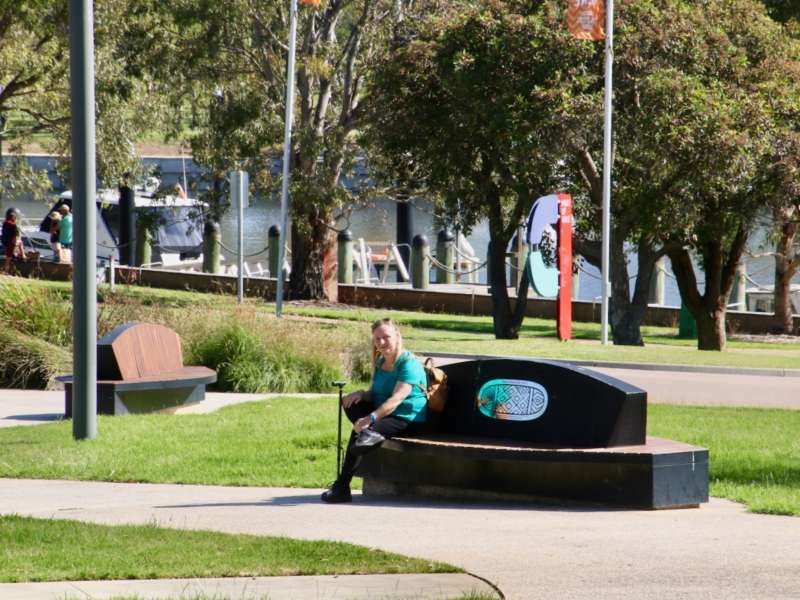 A person in a green shirt sits on a black bench in a park, surrounded by trees and a waterfront in the background.