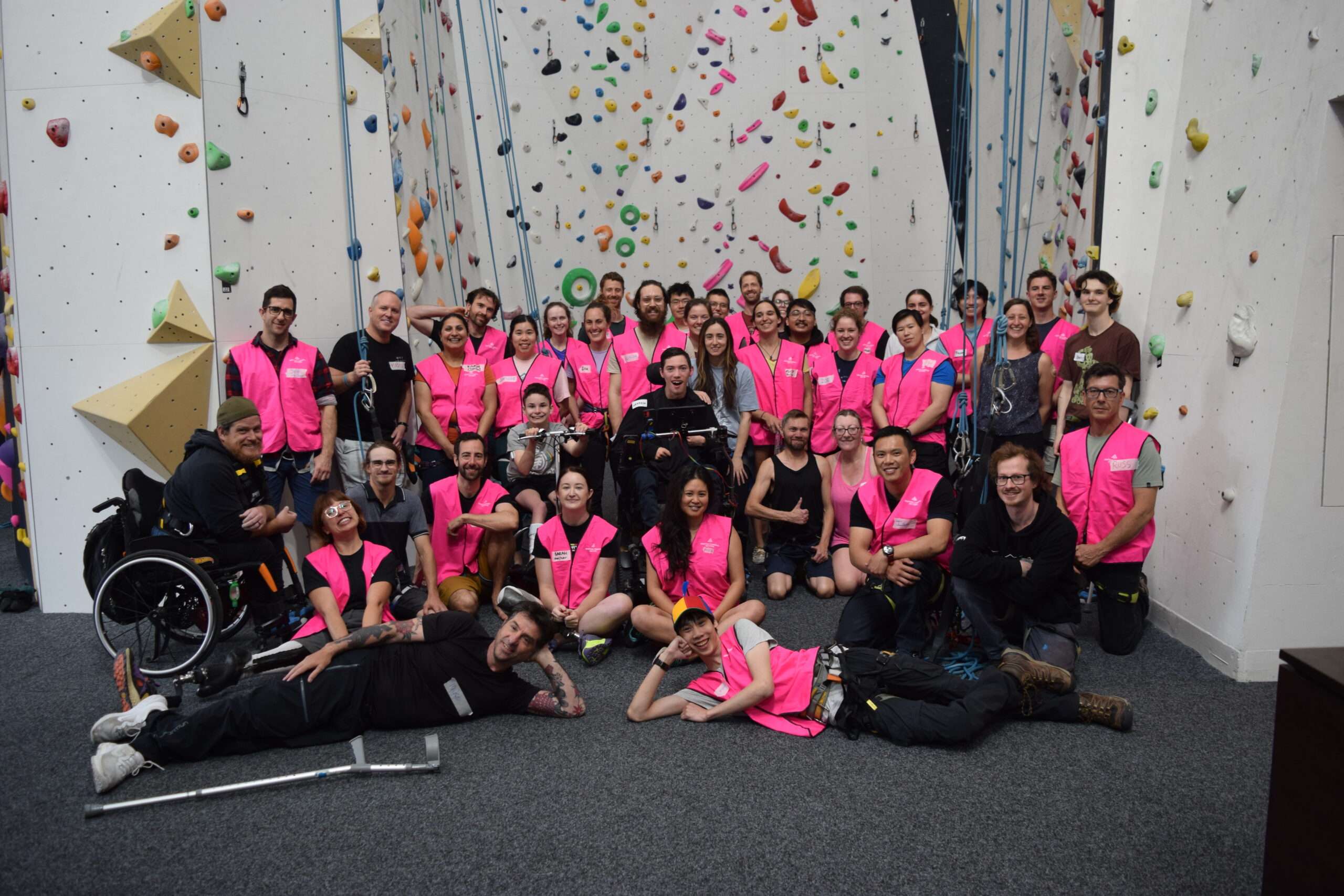 A large group of climbers wearing pink vests poses together in a climbing gym with colorful walls in the background.