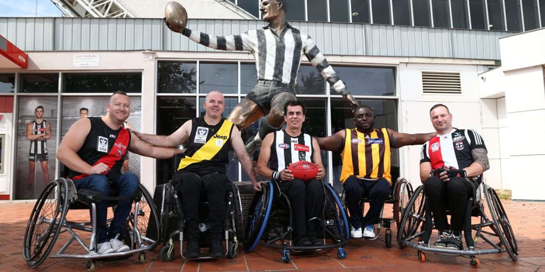 Five wheelchair athletes pose joyfully in front of a sports statue, showcasing team jerseys from various clubs, celebrating inclusivity.