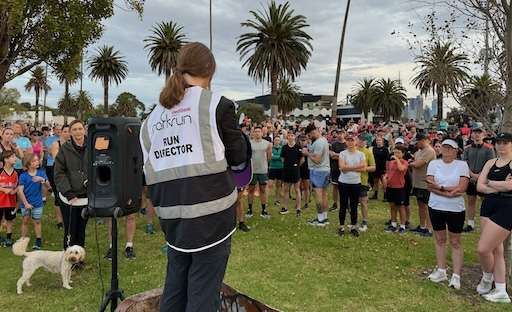 A run director speaks to a large gathering of runners in a park, with palm trees and city skyline in the background. A dog is nearby.