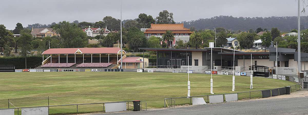 A large oval with green grass and goal posts with a red brick grand stand.