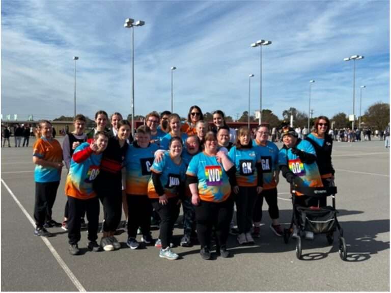 Photo of a group of men and women, on a netball court in Blue rainbow coloured t shirts smiling at the camera