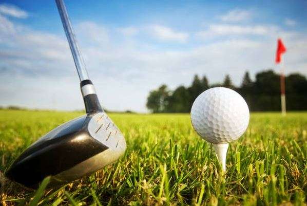 A golf club poised to strike a white ball teed on lush green grass, with a red flag in the background under a bright blue sky.