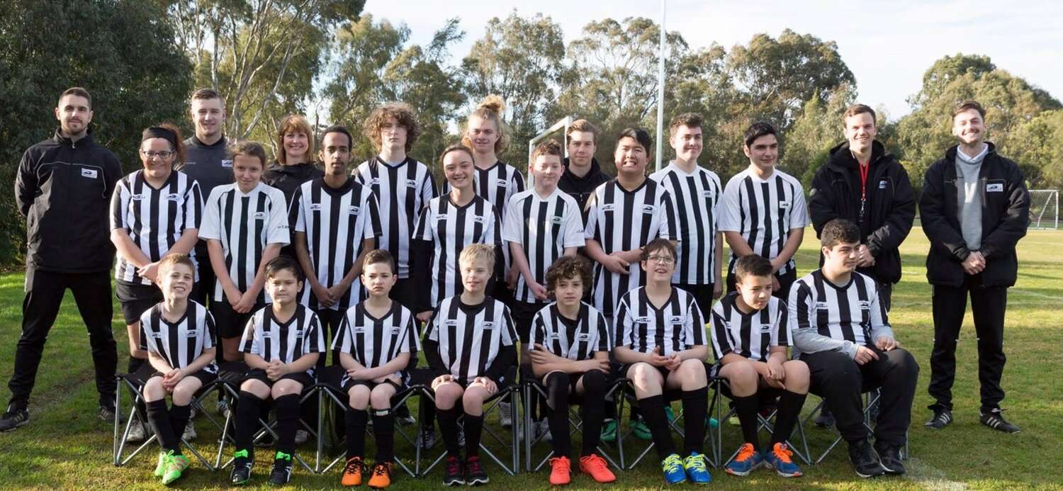 A youth soccer team poses together in striped jerseys, with coaches and staff, in a sunny outdoor setting.