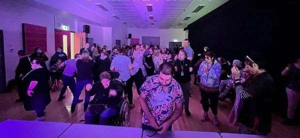 A lively crowd dances in a dimly lit hall adorned with purple lighting, showcasing joyful interactions among attendees.