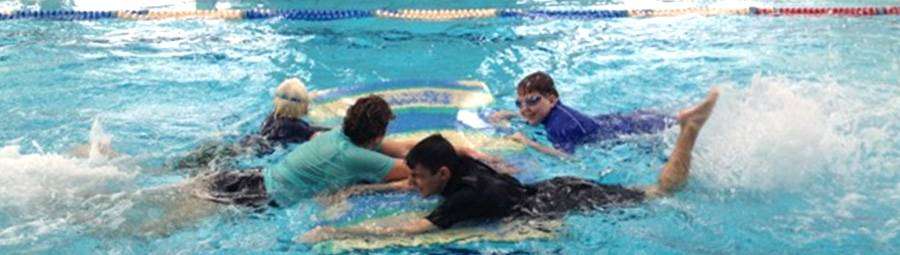 Children enjoying a swimming lesson, splashing in the water as they gather around a floating surfboard in a pool.