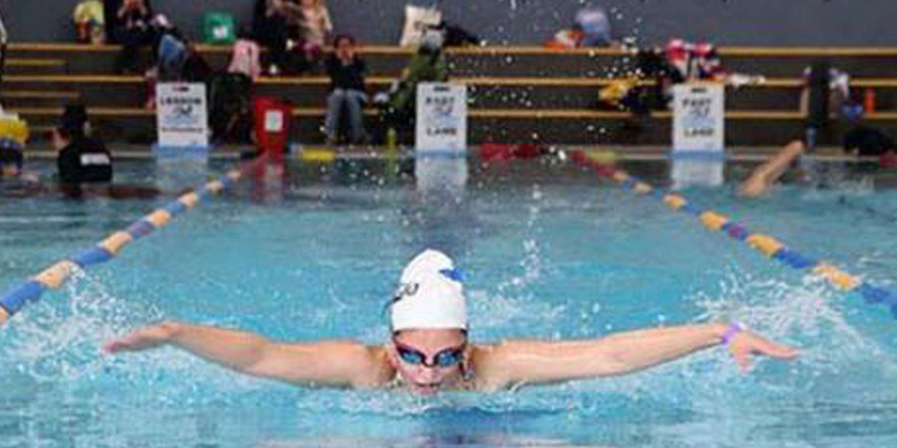 A young female swimmer in a white cap and goggles performs a butterfly stroke in a competitive swimming pool, with spectators in the background.