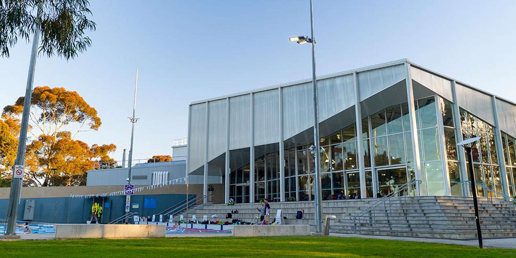 Modern glass-walled building with a peaked roof, surrounded by greenery and stairs leading to an outdoor area.