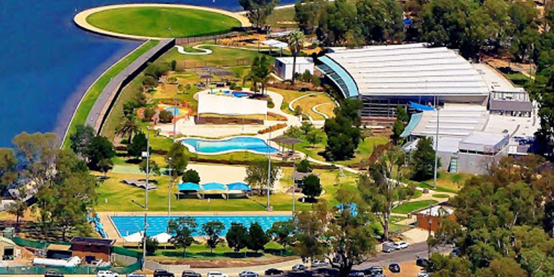 Aerial view of a recreational complex featuring pools, landscaped greenery, and a large building near a lake.