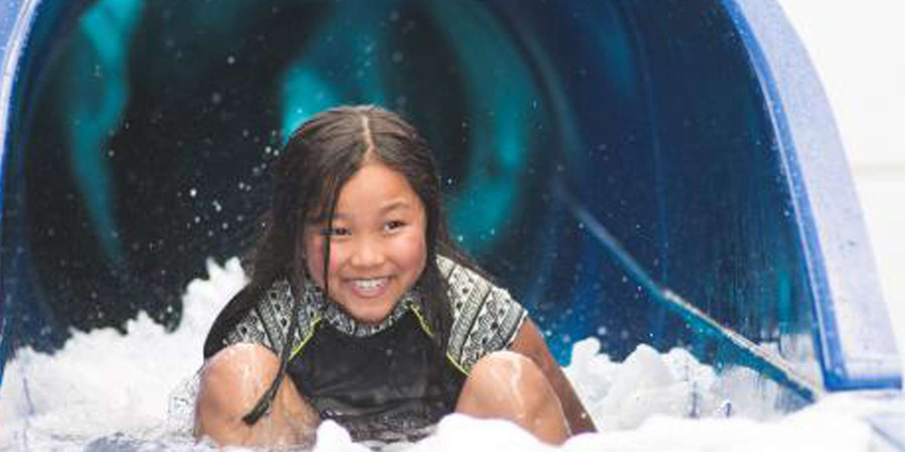 A child prepares to slide down a colorful water slide, surrounded by splashing water and a vibrant blue tunnel.
