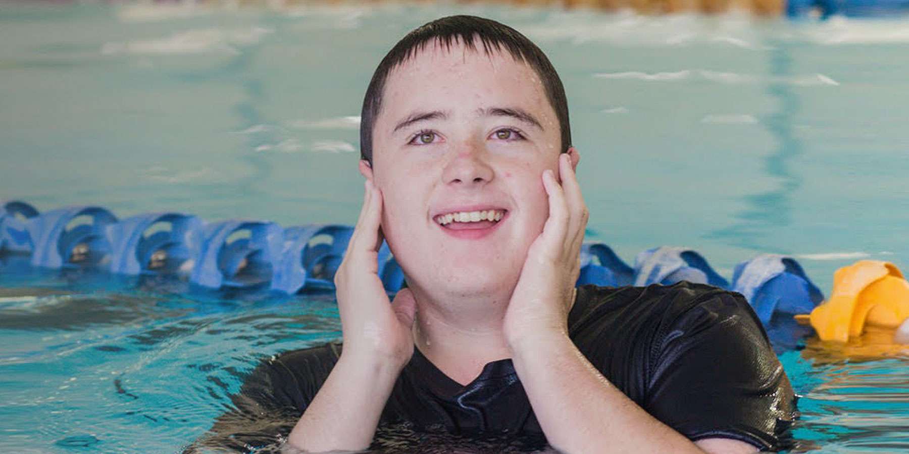 Image of a boy wearing a black top with his hands around his face smiling whilst swimming in an indoor swimming pool