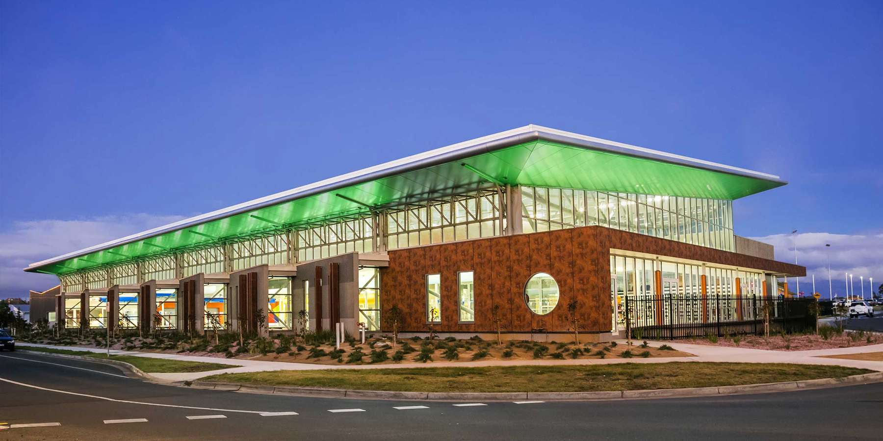Modern building with large glass windows and a green roof, illuminated at dusk, located at a street corner.
