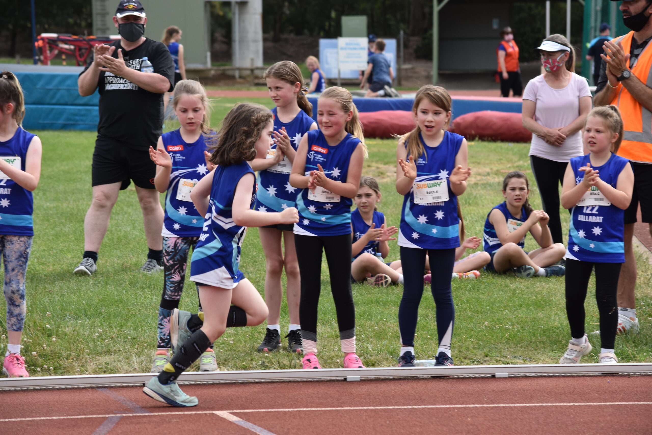 A young girl with a prosthetic leg runs on a track while a group of cheering peers in blue uniforms applaud her achievement.