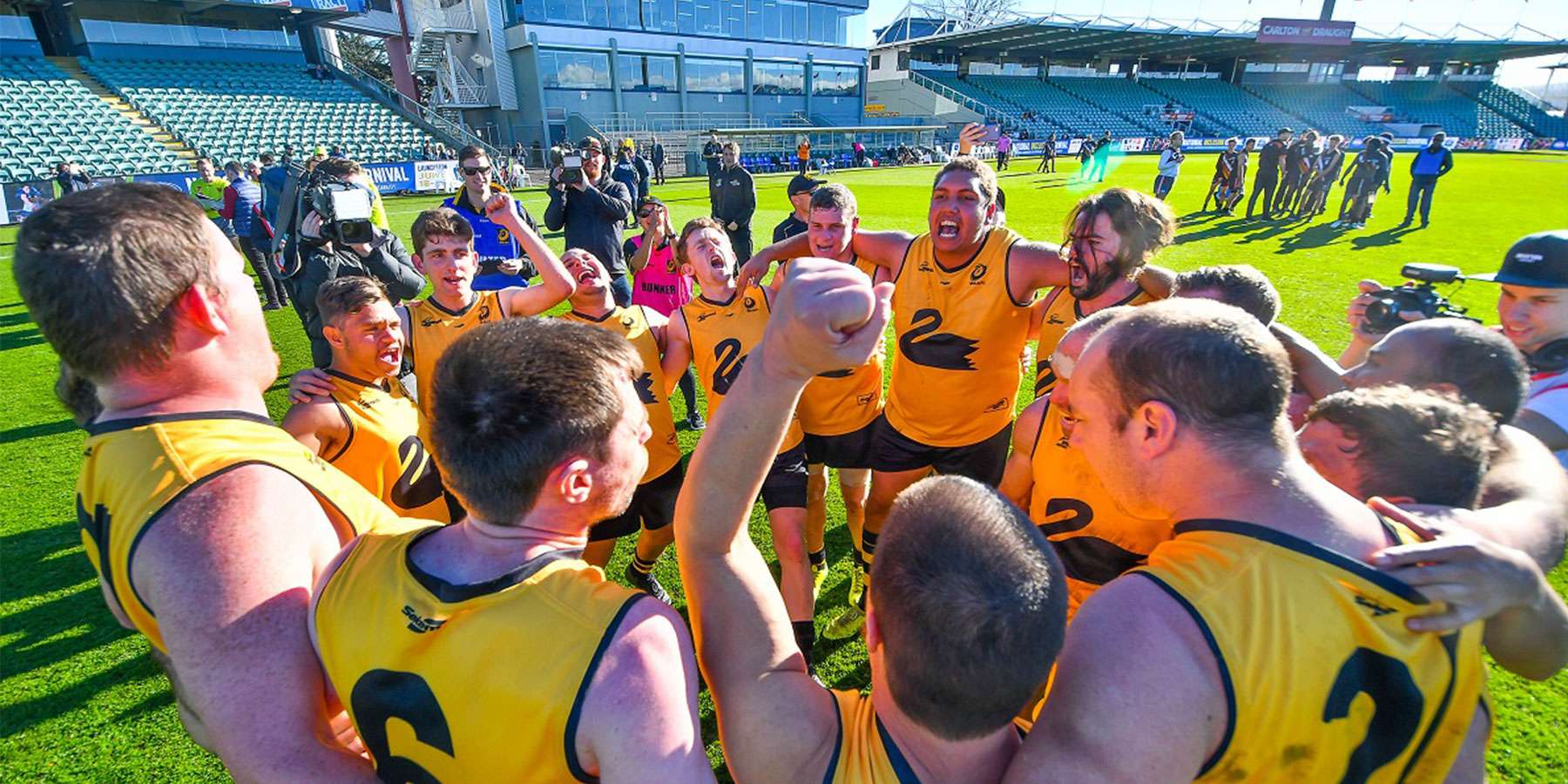 A team of athletes in yellow jerseys huddle together, celebrating and strategizing on a sunny field after a game.