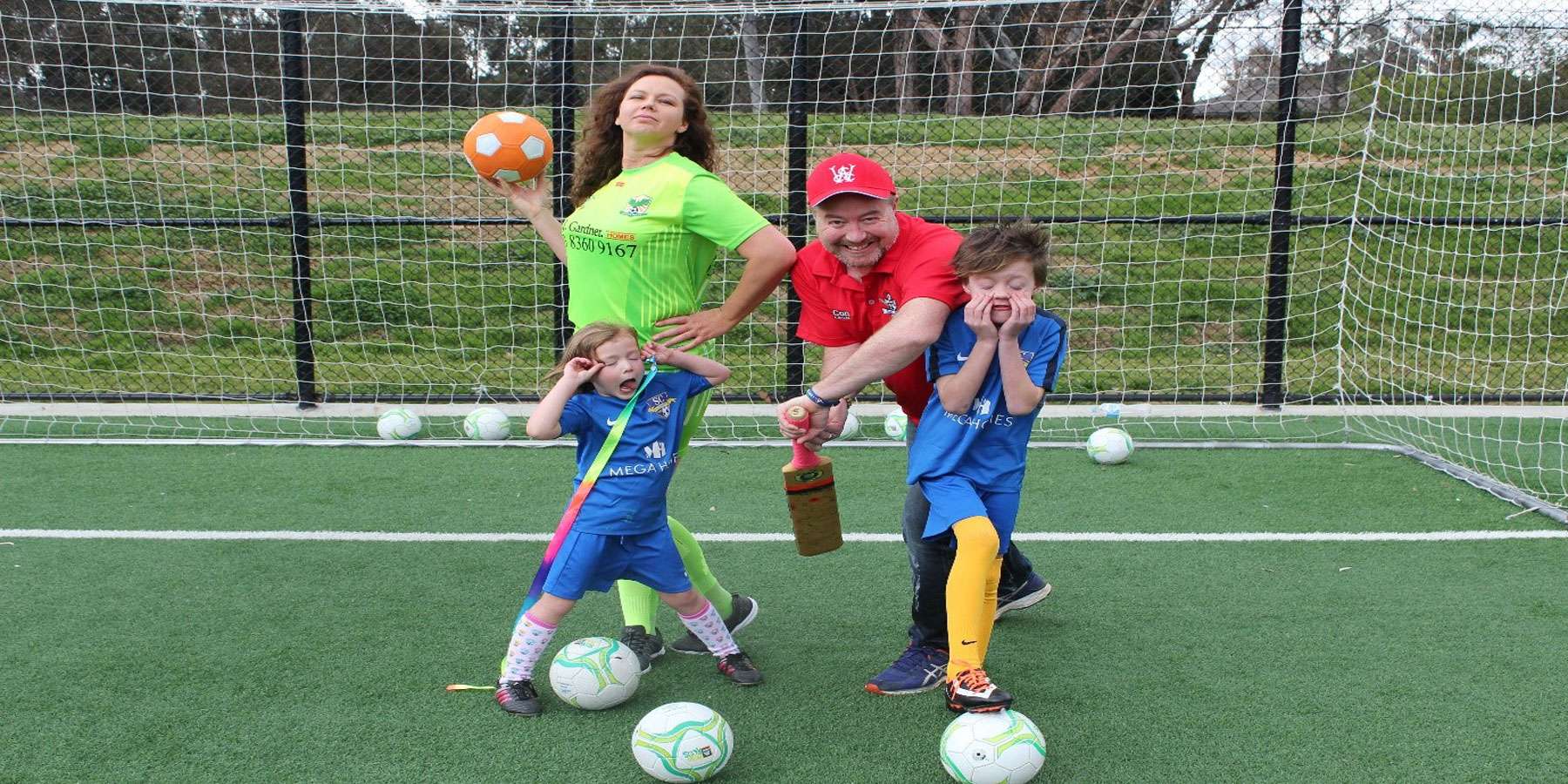 A playful scene on a soccer field features a woman in a bright green shirt, a man in a red cap, and two kids posing with soccer balls.