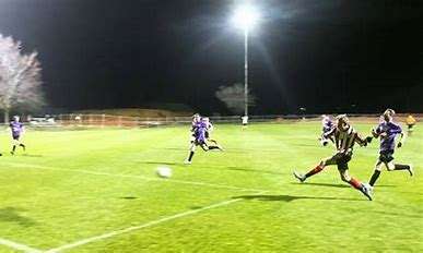 A soccer player in a striped jersey kicks the ball while players in purple chase after it under bright stadium lights at night.