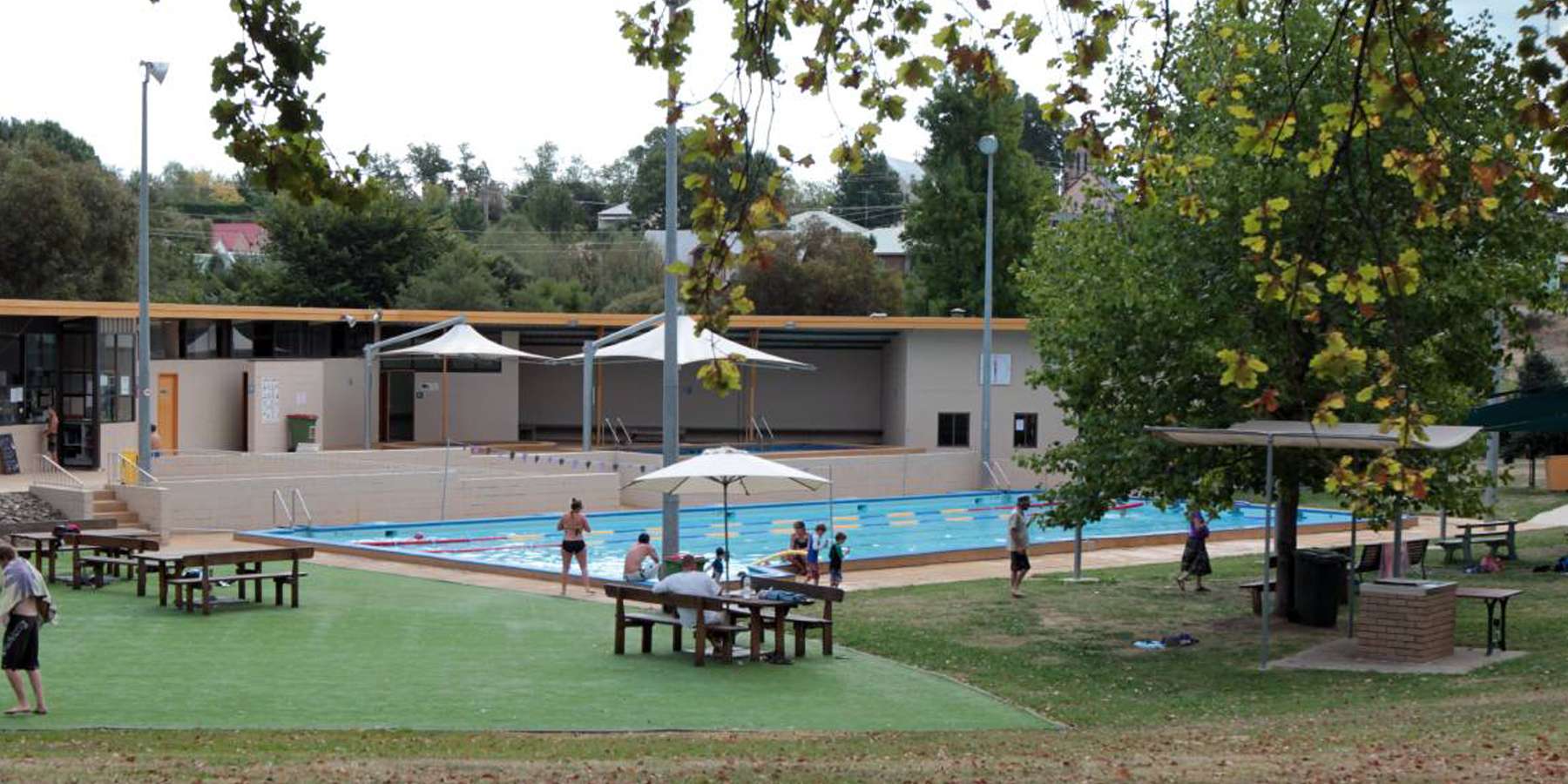 A swimming pool area with children playing, surrounded by greenery, picnic tables, and shaded seating under umbrellas.