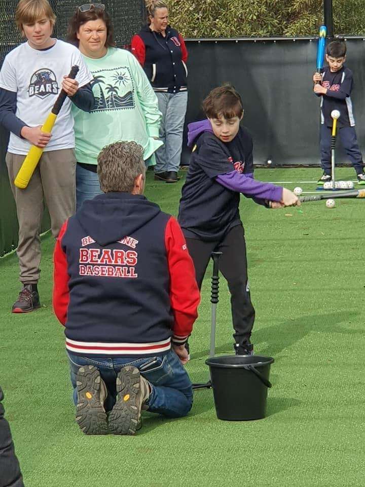 A child practices baseball hitting with a coach's guidance, surrounded by other players on a grassy field at a training session.
