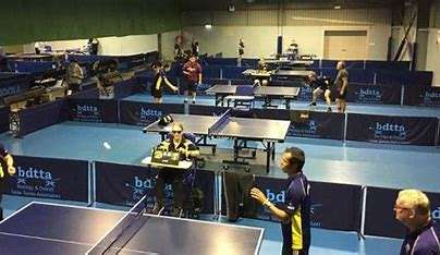 Image of mutiple people playing table tennis in a purpose built table tennis space. There are 5 tables in the image, and players are wearing blue and yellow uniforms playing table tennis