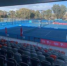 View of a tennis court with empty bleachers, surrounded by greenery and blue fencing, set for a tournament atmosphere.