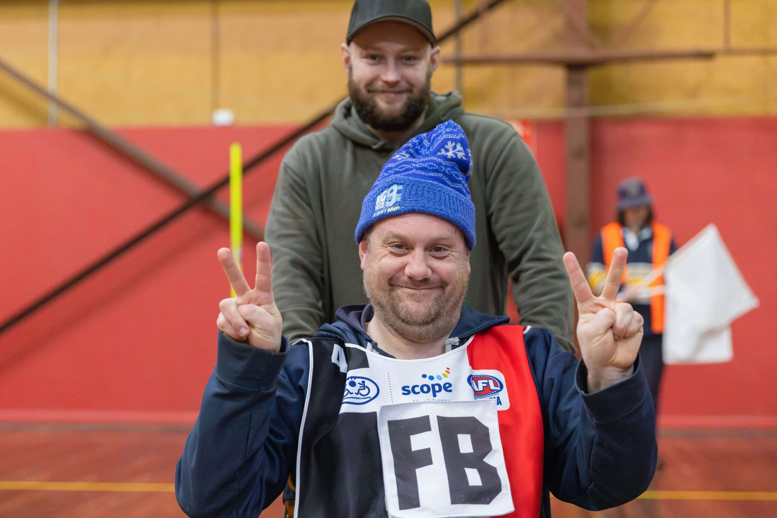 A man in a wheelchair is wearing a sports uniform and beanie. He is smiling and holding his hands up. Another man stands behind him and is also smiling