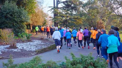 A large group of runners in colorful athletic wear jogs along a tree-lined path in a park, surrounded by greenery and remnants of snow.