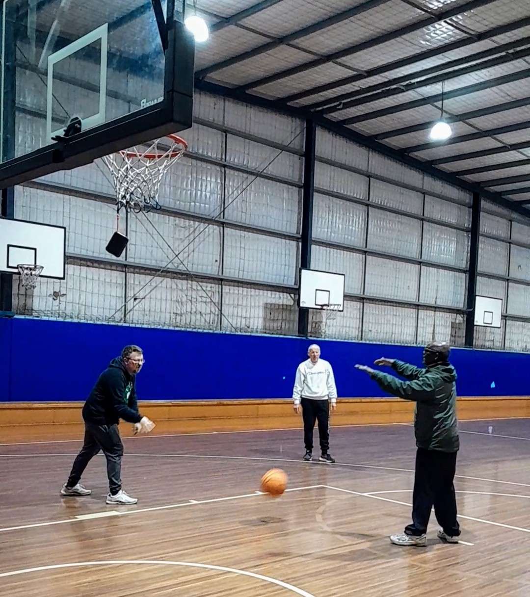 Two people practice basketball in a gym while another watches, with hoops in the background and a basketball on the court.