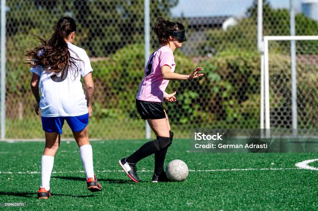 Photo showing two women playing soccer, one woman in blue shorts, white long socks and white t shirt, second women in a pink shirt, black shorts, long black socks kicking a soccer ball towards the goals