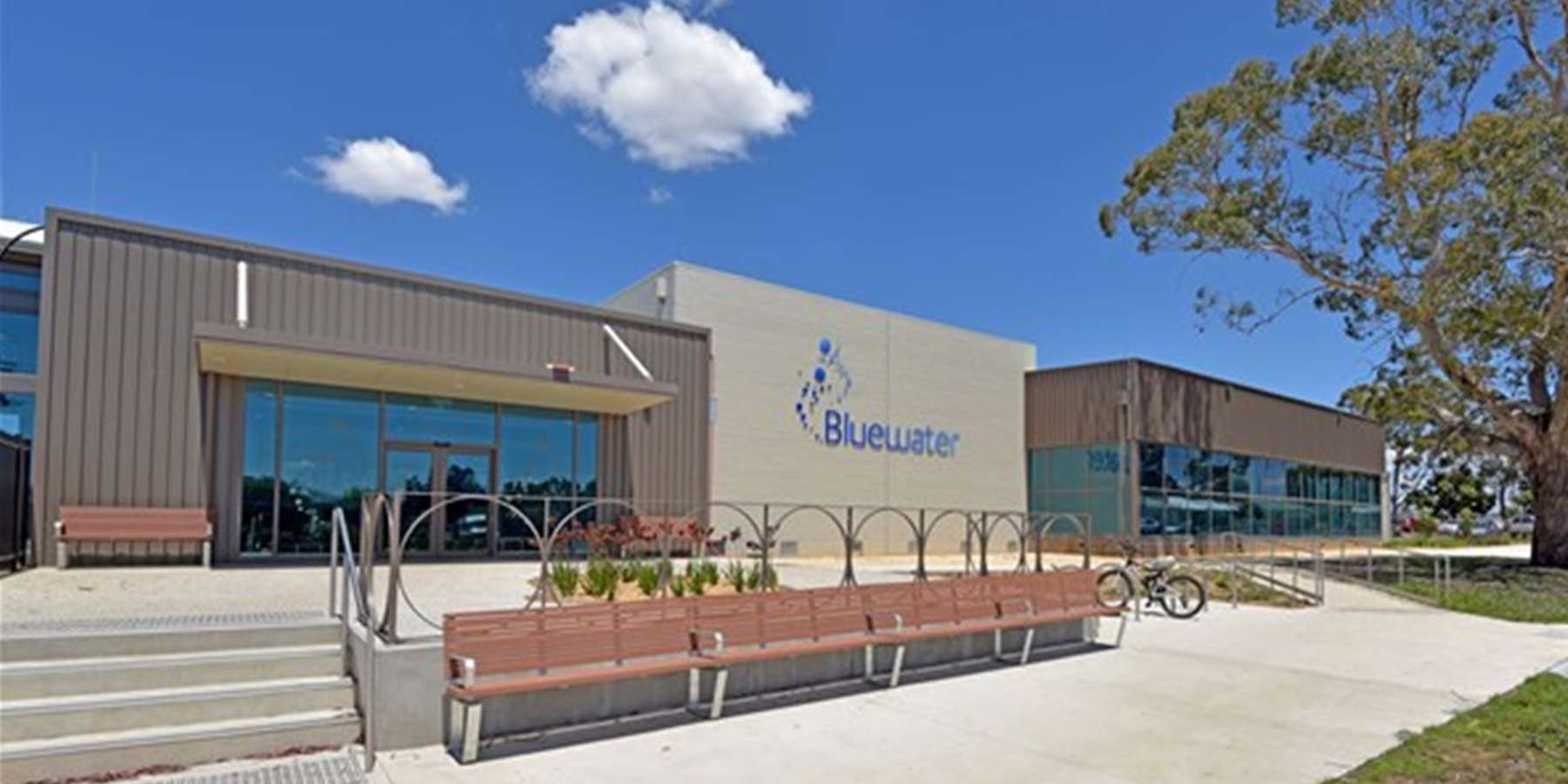 Modern building exterior with a logo, benches, and bike rack under a blue sky with fluffy clouds.