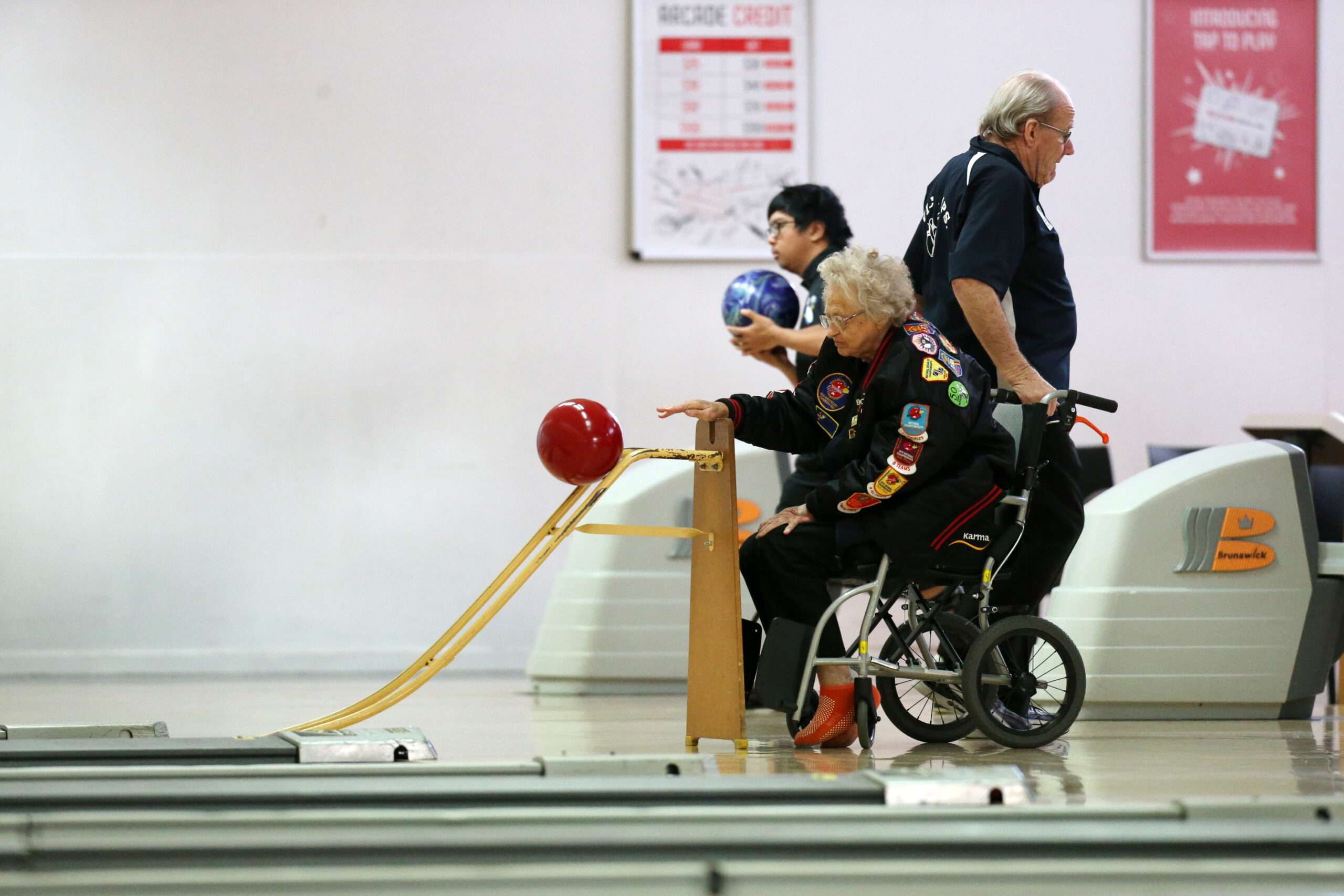 Photo of tenpin bowling lanes with lady in a wheelchair using the accessibility ramp to roll the red bowling ball down the lane. Two other bowlers stand close by.