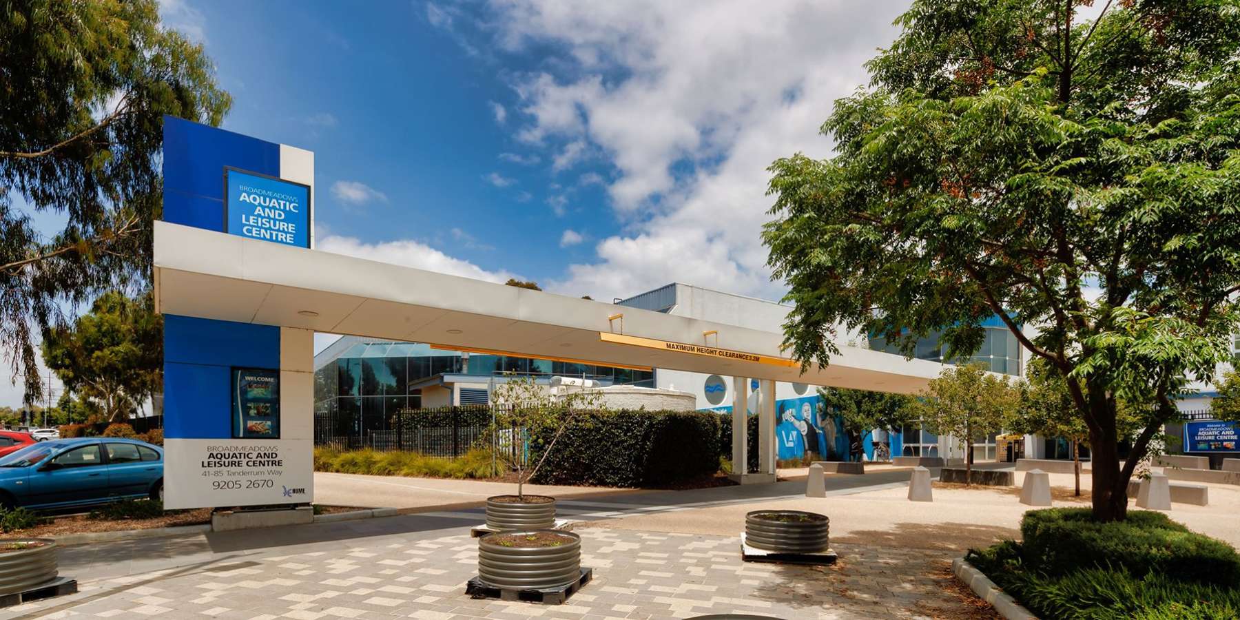 Modern building with blue accents, surrounded by greenery and featuring a pedestrian pathway under a bright, partly cloudy sky.