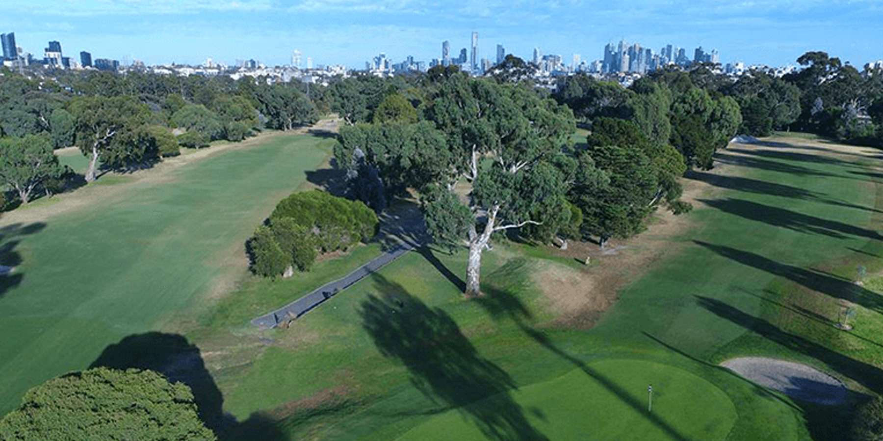 Aerial view of a lush golf course with manicured fairways, large trees, and shadows casting over the green landscape under a clear blue sky.