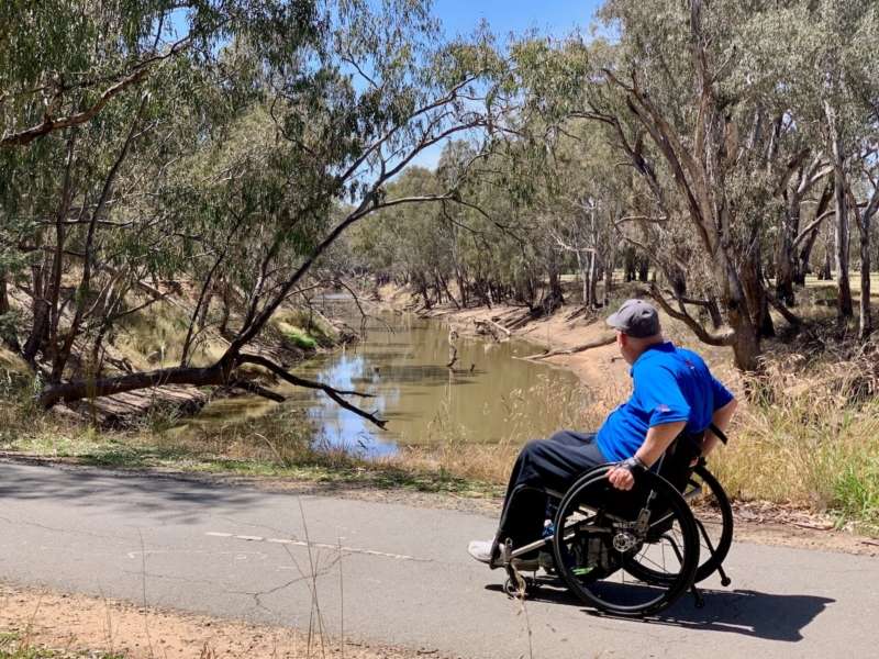 A man in a blue t shirts and black pants in a wheelchair next to the Campaspe River