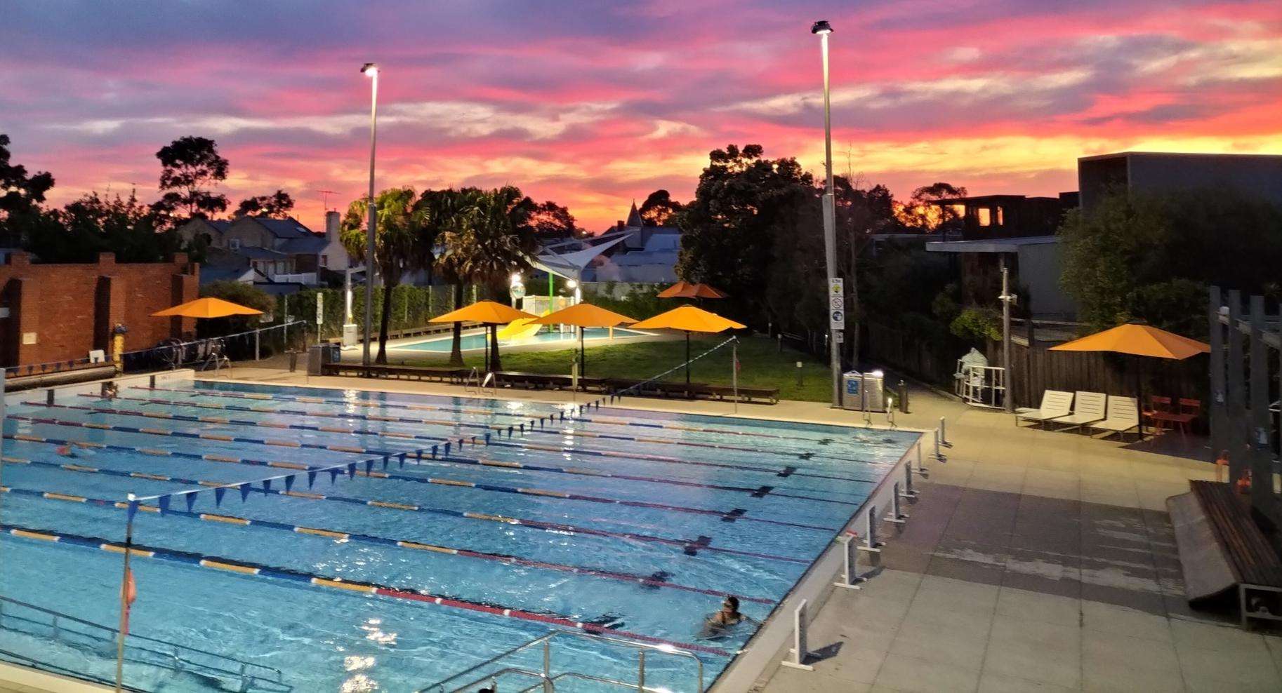 A large inground pool surrounded by green trees and grass and a brown brick changeroom in the background.