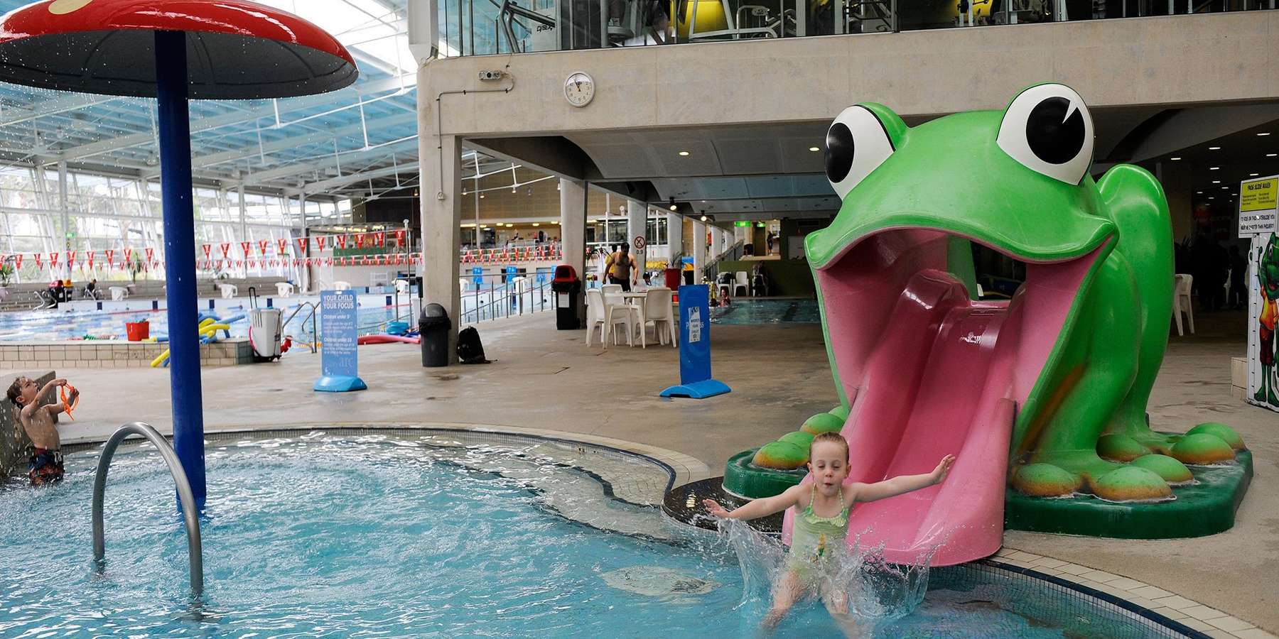 A colorful indoor water park featuring a large frog slide, a child playing in the pool, and water features, creating a fun atmosphere.