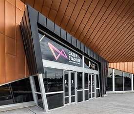 Image displays the Casey Stadium building. The building is black, grey and orange in colour with the purple and pink Casey Stadium logo on the window