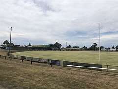 Wide view of a sports oval with a grassy field, surrounding fence, and a modest grandstand under a cloudy sky.
